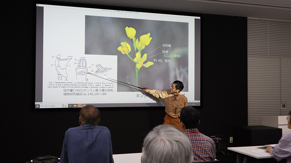 スライド投影したキンランの資料について説明する、オレンジ色のサイトウエアを着たオンライン カジノ 大 負け社員